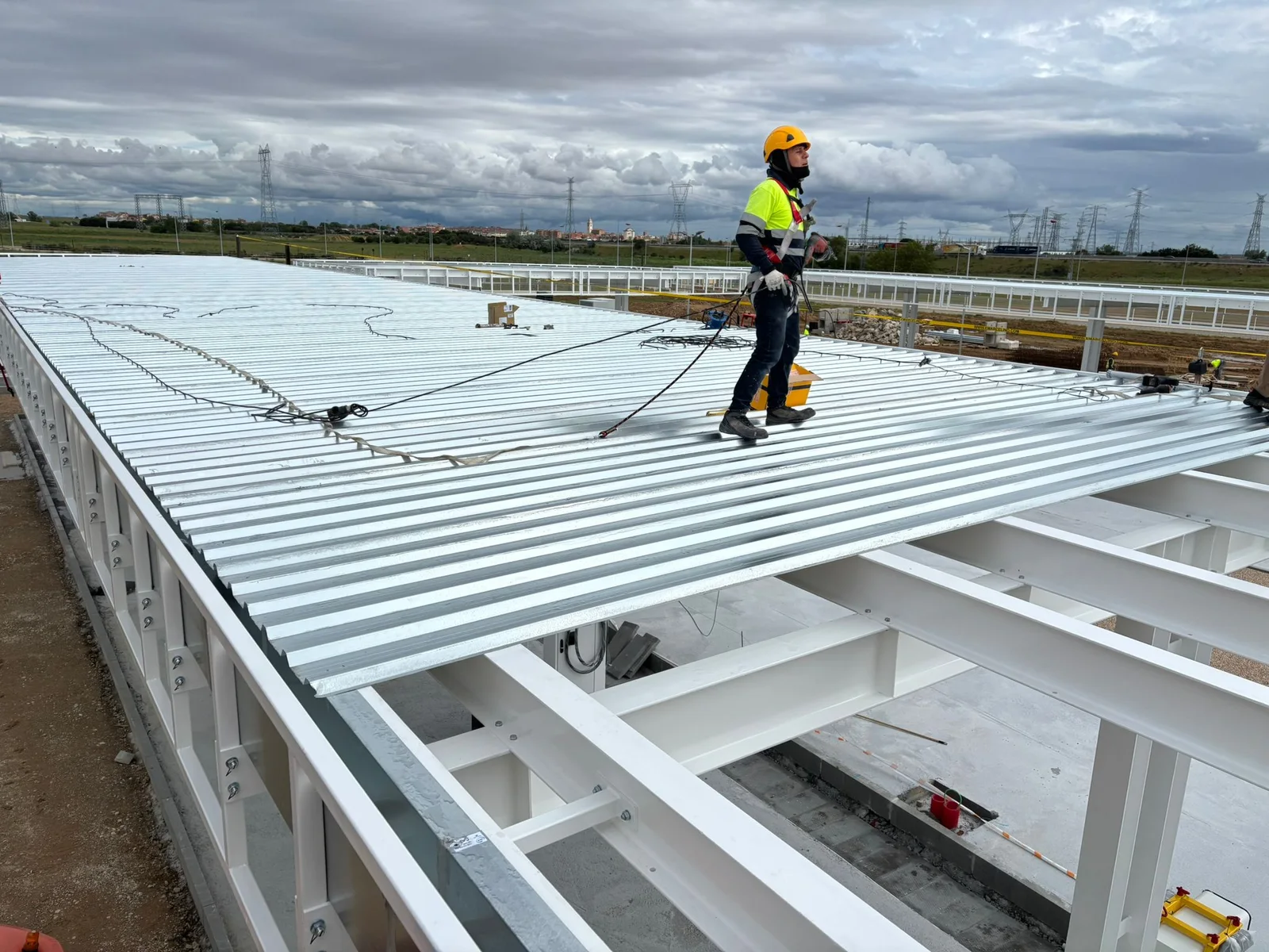 Montagem de cobertura metálica sobre estrutura de aço, com trabalhador equipado com capacete e arnês
